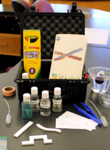 A close-up photo of the elements of the Chem-O-Meter kit showing colored pencils, a work booklet, vials of liquid, and test paper.