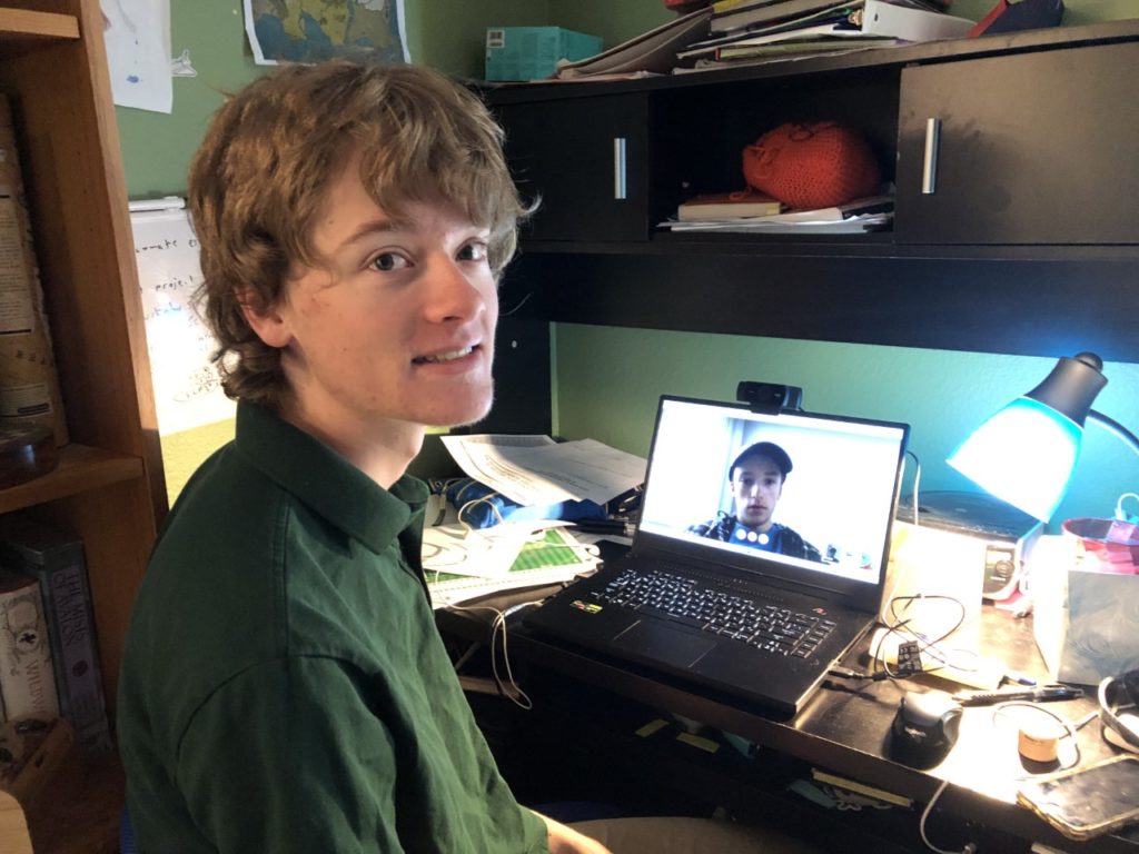 An 18-year-old smiles at the camera in front of a laptop. He is video calling his partners for the science fair