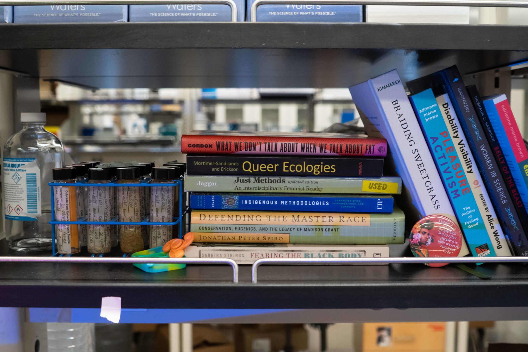 Books about Queer Ecology, and other Diversity, Equity and Inclusion concepts stacked on a shelf in a lab next to glasses of scientific tests.